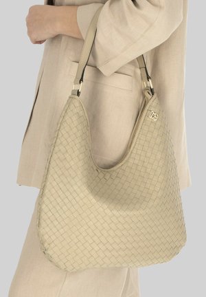 Beżowa torebka z plecionej skóry z złotym logo, trzymana przez osobę ubraną w dopasowany beżowy garnitur z rękawami do łokcia.