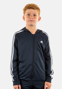 Felpa con zip color navy, con strisce bianche laterali, realizzata in tessuto morbido. Presenta una tasca frontale e un cappuccio, con un piccolo logo sul petto sinistro.