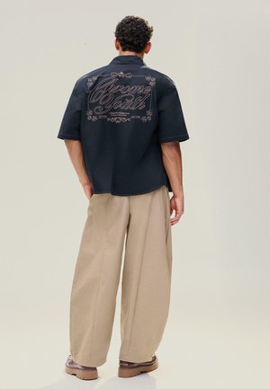 Ein Mann steht mit dem Rücken zur Kamera, trägt ein schwarzes kurzärmeliges Hemd mit der Aufschrift "Chrome South" und beige, weit geschnittene Hosen mit braunen Schuhen.