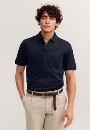 Junger Mann mit lockigem Haar, der ein schwarzes kurzärmeliges Poloshirt, beige Hosen und einen braunen geflochtenen Gürtel trägt, steht mit den Händen in den Taschen.