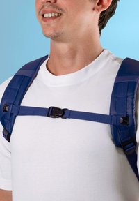 Justerbar blå ryggsekkreim med spenne, båret over en hvit skjorte, med polstret materiale for komfort og sikker passform.