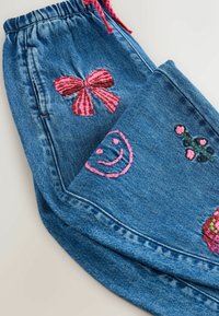 Joggers in denim con vita regolabile, caratterizzati da ricami fucsia: un fiocco, una faccina sorridente e fiori su un tessuto blu testurizzato.