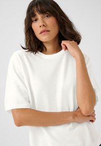 Pull blanc à manches courtes avec un col rond côtelé. Texture douce avec une coupe décontractée, présentant des bords légèrement roulés sur les manches.