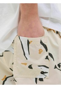 Shorts beiges avec un motif de formes noires, blanches et moutarde. Dotés d'une poche latérale avec un détail de bouton noir. Texture lisse.
