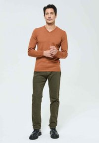 Un pull brun en V, un pantalon vert slim et des chaussures noires. Le pull est texturé ; le pantalon a une finition lisse. Tenue décontractée sur un fond neutre.