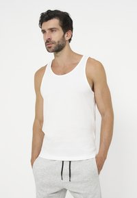 Homme aux cheveux foncés et à la barbe, portant un débardeur blanc et un pantalon de jogging gris clair, les mains dans les poches, regardant sur le côté.