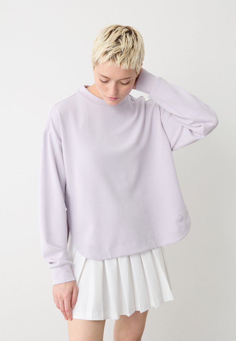 Felpa oversized color lavanda chiaro con maniche lunghe, orlo arrotondato e discreto dettaglio del logo. Indossata con una gonna bianca plissettata.