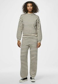 Sweter i spodnie w paski w kremowym i czarnym kolorze. Sweter ma wysoki kołnierz i luźne rękawy; spodnie są szerokie. Czarne buty.