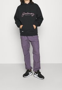 Černá mikina s kapucí s nápisem "Grimes Mud City" v mnohobarevném písmu, v kombinaci s fialovými cargo kalhotami a černými sportovními botami.