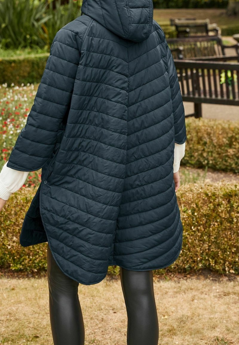 Personne portant un manteau matelassé à capuche bleu foncé avec des manches trois-quarts, debout à l'extérieur près de haies taillées et de bancs de parc.