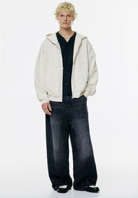 Cremefarbene Kapuzensweatshirt mit lässigem Schnitt aus strukturiertem Stoff. Dunkelblaues Hemd darunter, kombiniert mit lässigen schwarzen Jeans und schwarzen Schuhen.