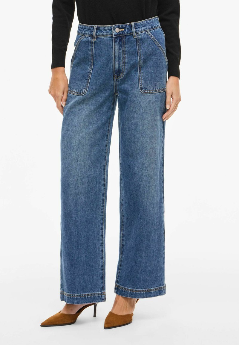 Vide bukser i denim med middels blå farge, som har høy midje, store fremre lommer og kontrastsøm. Kombinert med brune hæler.