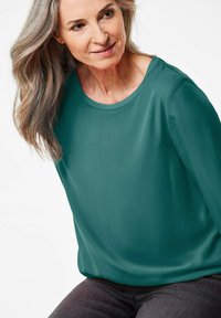 Blusa lunga teal a maniche lunghe con scollatura rotonda, realizzata in tessuto morbido. Presenta una vestibilità ampia e una tasca laterale discreta. Indossata con jeans scuri.
