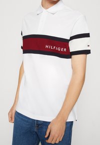 Valkoinen polo-paita lyhyillä hihoilla, jossa on punaisia ja laivastonsinisiä vaakaraitoja sekä "HILFIGER"-logo. Valmistettu sileästä puuvillasekoitekankaasta.
