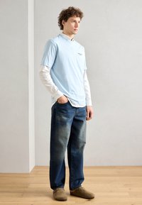 Fakék, rövid ujjú póló, fehér, hosszú ujjú ing fölé rétegezve, amelyet elhasználódott kék farmernadrággal és barna slip-on cipőkkel párosítanak.