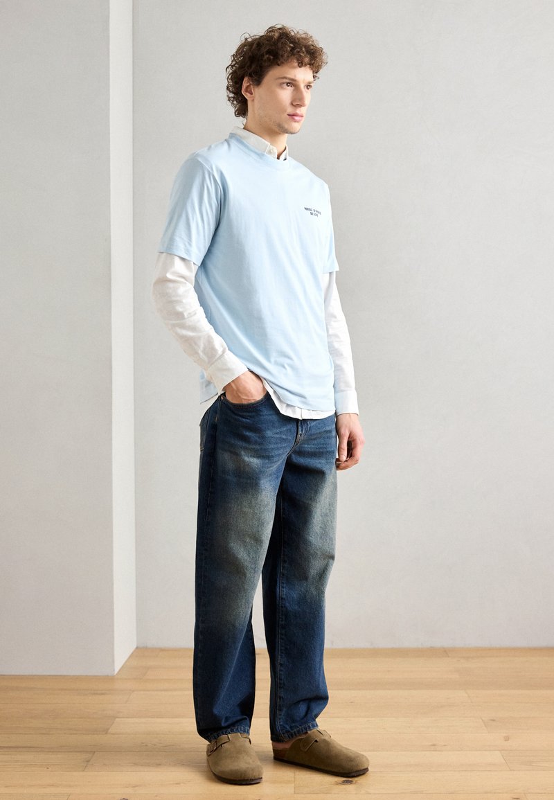 Hellblaues kurzärmliges T-Shirt, das über einem weißen langärmligen Hemd getragen wird, kombiniert mit ausgewaschenen blauen Jeans und braunen Slippern.