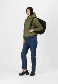 Olivengrønn pufferjakke med hette, svart quiltet ryggsekk, blå jeans og svarte loafers. Jakken har en frontglidelås og ribbestrikkede mansjetter.