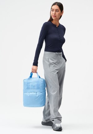 Frau in marineblauem Langarmshirt und grauer Hose hält einen hellblauen Rucksack an den Griffen und schaut seitlich, während sie vor weißem Hintergrund steht.