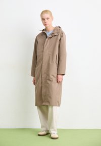 Beige vattentät kappa med huva, lång design, sidofickor och en slät textur, kombinerad med krämfärgade byxor och ljusa skor.