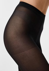 Gros plan sur la hanche et le haut de la cuisse d'une personne portant des collants noirs transparents sur un fond blanc uni.