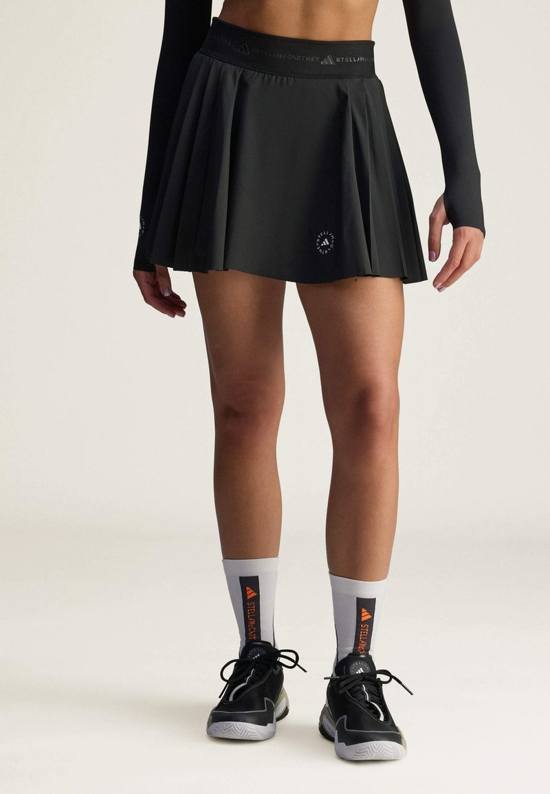 Falda negra con pliegues y cinturilla ancha, combinada con zapatillas negras y blancas y calcetines grises con acentos naranjas y marca.