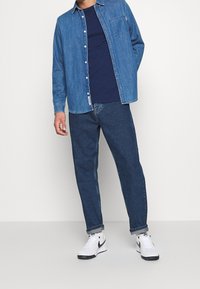 Camisa de mezclilla de botones en azul claro sobre una camiseta navy, combinada con jeans azul oscuro y zapatillas blancas con detalles en negro.