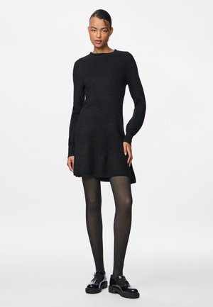 Abito lavorato a maglia nero con maniche lunghe, scollatura rotonda e orlo svasato. Indossato con collant neri e scarpe nere lucide.