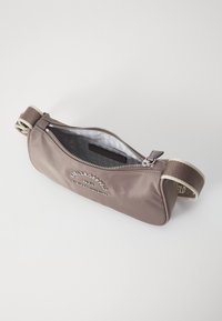 Brun tygväska med ett dragkedjelock, rund form och en vävd rem. Har en präglad logotyp och en slät, glansig yta. Interiören är grå.