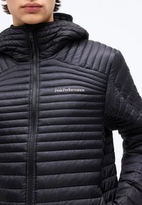 Κοντινό πλάνο ενός ατόμου που φοράει μαύρο καπιτονέ μπουφάν με φερμουάρ και κουκούλα, με το λευκό λογότυπο "Peak Performance" στο στήθος.