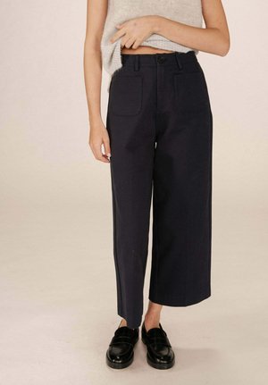 Pantalons larges marins en tissu texturé, dotés de deux poches avant, d'une fermeture à bouton à l'avant et d'un ourlet droit. Mocassins noirs portés.