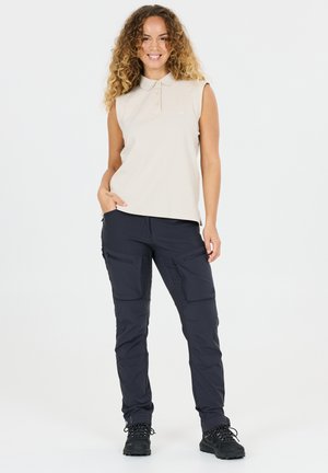 Femme souriante aux cheveux bouclés portant un polo beige sans manches, un pantalon cargo noir et des chaussures de randonnée noires, debout avec une main dans la poche.