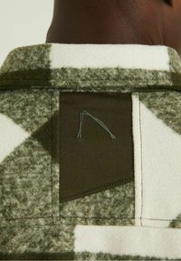 Gros plan sur le col d'une veste en laine à carreaux verts et blancs, avec un patch en tissu vert olive présentant un motif minimaliste cousu.