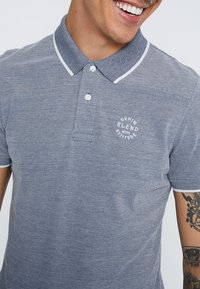 Marineblå polo shirt med stribet krave, korte ærmer og logo på forsiden. Lavet af et tekstureret stof med knaplukning og kontrastsyning.