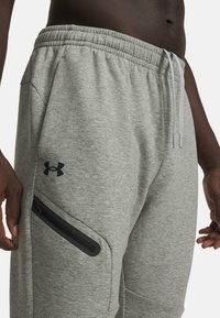 Graue Jogginghose aus weichem Stoff mit elastischem Bund, Kordelzug und Reißverschlusstasche am linken Oberschenkel. Schwarzes Logo-Akzent.