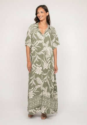 Femme souriante debout, portant une robe maxi vert olive avec des motifs floraux et feuillages blancs et des manches courtes.