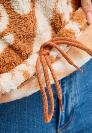Braune und weiße Fleecejacke mit einem braunen Kordelzug, der zu einer Schleife gebunden ist, über blauen Jeans, gehalten von Händen mit nude-farbenem Nagellack.