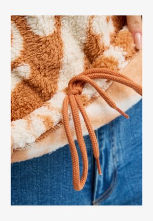 Braune und weiße Fleecejacke mit einem braunen Kordelzug, der zu einer Schleife gebunden ist, über blauen Jeans, gehalten von Händen mit nude-farbenem Nagellack.