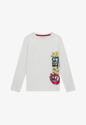 Chemise blanche à manches longues avec un grand texte vertical coloré "GUESS" sur le côté droit en lettres rouges, jaunes, bleues et noires.