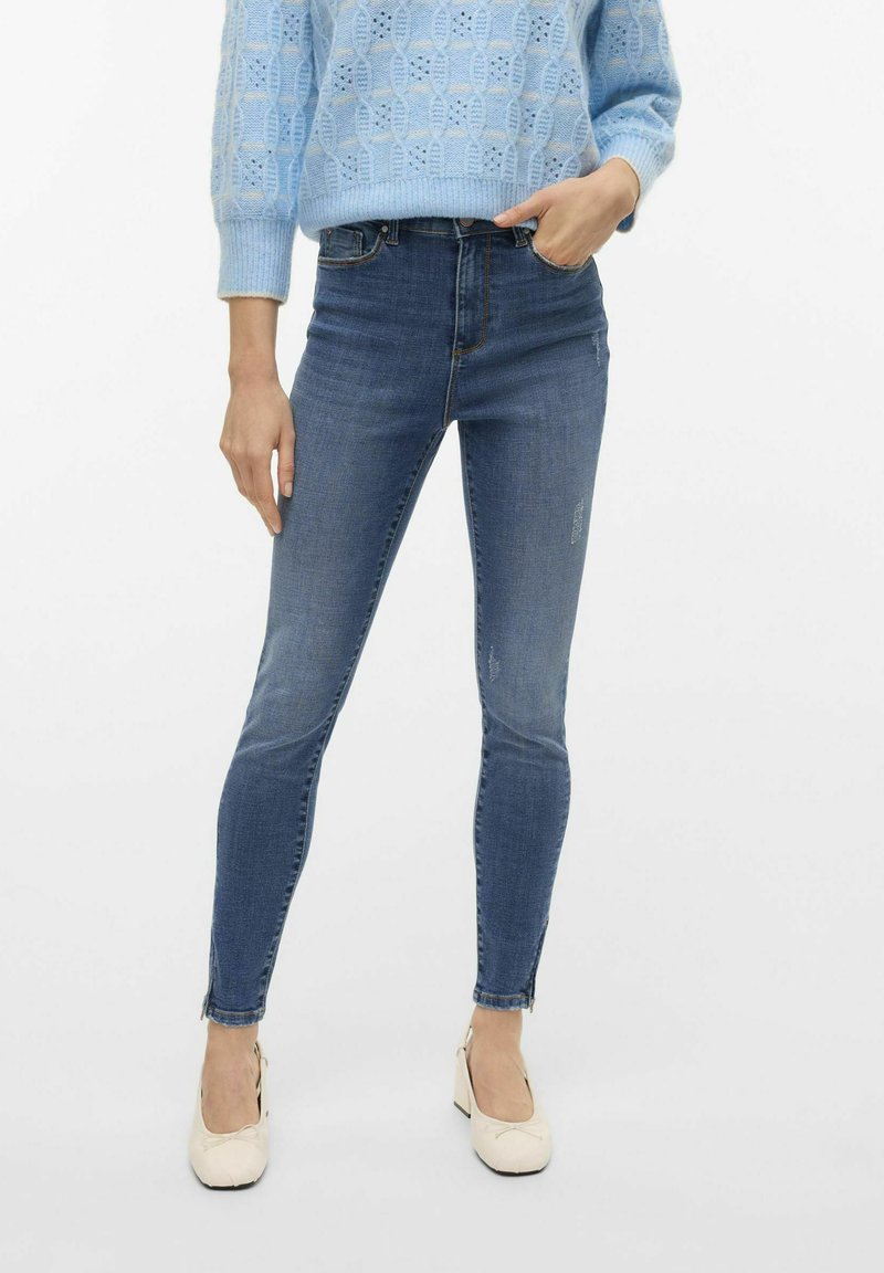 Jeans skinny en denim bleu avec une taille mi-haute, un léger délavage et des accents de fermeture éclair au niveau de la cheville, associés à un pull en tricot bleu clair.