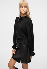 Νεαρή γυναίκα με ξανθά μαλλιά που φοράει μαύρο μπλουζάκι με μακριά μανίκια και δαντελένιες λεπτομέρειες, καθώς και μαύρη δερμάτινη φούστα με σχίσιμο μπροστά, ποζάροντας πλάγια.