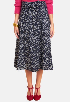 Woman wearing navy blue midi skirt with white floral print and bow belt, red T-strap shoes, pink sweater, and gold bracelets.