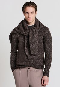 Braun gefleckter Strickpullover mit geripptem Rundhalsausschnitt und �übergroßem Kragen. Verknoteter Schal über die Schultern drapiert. Hellbraune Hose.