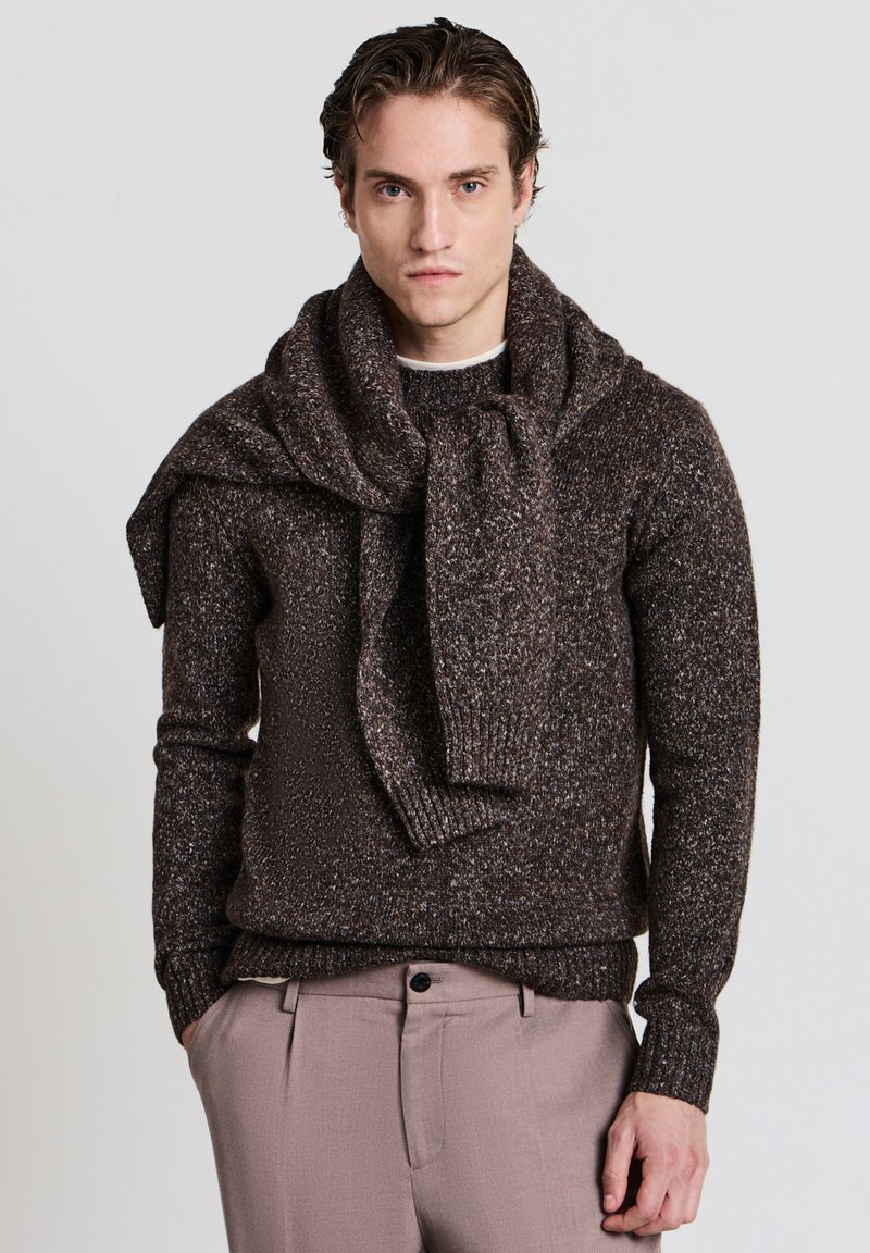 Braun gefleckter Strickpullover mit geripptem Rundhalsausschnitt und übergroßem Kragen. Verknoteter Schal über die Schultern drapiert. Hellbraune Hose.