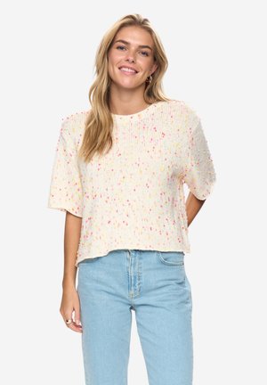 Smilende kvinde med langt blondt hår iført en cremefarvet, strikket kortærmet sweater med lyserøde og gule pletter og lyseblå jeans.