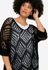 Schwarzer offener Strick-Poncho mit geometrischem Diamantmuster, weiten Ärmeln und rundem Ausschnitt über einem weißen Shirt.