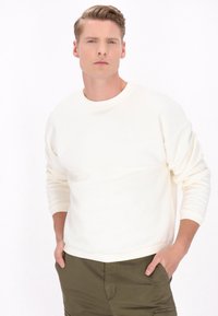 Sweatshirt crème avec un col rond, des poignets côtelés et une texture lisse ; présente un logo subtil sur la poitrine. Associé à un pantalon vert olive.