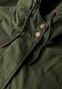 Veste à capuche verte en tissu texturé, dotée de boutons-pression en bronze et de cordons avec embouts en métal. Vue en gros plan des détails du design.