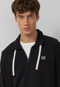 Czarna bluza zapinana na zamek z białymi troczkami, z naszywką z logo i teksturowanym kołnierzem. Nałożona na białą koszulkę.
