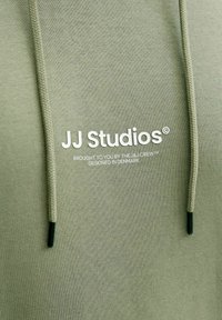Grön hoodie med en jämn yta, utrustad med dragsko och en logotyp tryckt i vitt: "JJ Studios" med ytterligare text nedanför.