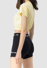 Žena v žlutém zkráceném tričku a černých cyklistických šortkách s vysokým pasem značky Champion stojí na jednotném světlém pozadí.